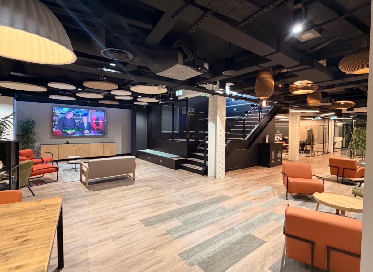 Interior of the Santander Work Café at Regina House, Cheapside. A warm open lounge with orange armchairs, soft wood floors, pendant lighting and a presentation screen at the front.